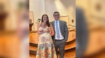 Un hombre en traje y una mujer con vestido estampado sostienen a un bebé vestido de blanco frente al altar de una iglesia.