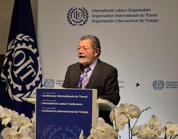 Hombre con barba y gafas habla en un podio frente a un logotipo y bandera de la OIT, con orquídeas blancas en primer plano