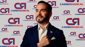 Hombre con barba en traje azul, camisa blanca, y pin de bandera colombiana, posando frente a un fondo repetido con los logos de Cambio Radical.