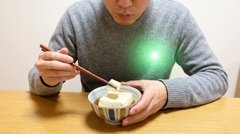 Un hombre sostiene un trozo de tofu con palillos sobre un tazón, mientras una luz verde ilumina la parte izquierda de su pecho.