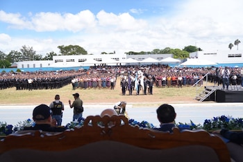 Gran multitud de personas, algunos con trajes tradicionales y otros con uniformes, en un campo. Se ven militares con banderas, fotógrafos y personal con chalecos de Policía