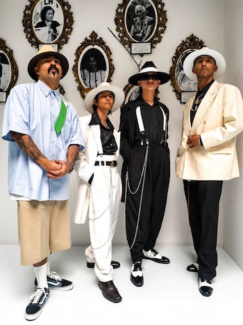 Cuatro hombres posan con sombreros fedora, trajes holgados blancos y negros, y pantalones cortos, frente a una pared blanca adornada con retratos en marcos dorados