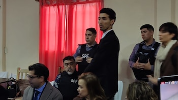 Un grupo de personas en un espacio interior con cortinas rojas. Varios hombres utilizan chalecos negros con la sigla SPF. Algunos hombres visten traje