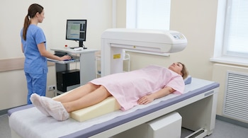 Una mujer de 40 años en bata rosa sobre una mesa de examen, con las piernas elevadas, bajo un equipo de densitometría ósea mientras una técnica monitoriza.