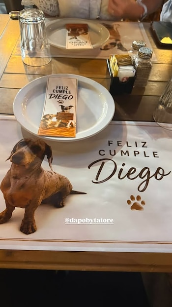 Mesa de madera con dos platos blancos, dos vasos, un salero, un pimentero, y dos individuales blancos con un perro salchicha y el texto 'Feliz Cumple Diego'