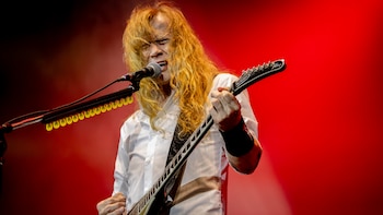 Primer plano de un guitarrista de pelo largo y rubio, con camisa blanca, cantando en un micrófono y tocando una guitarra eléctrica en un escenario con luz roja