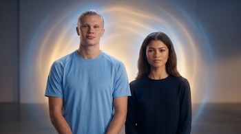 Erling Haaland de camiseta azul claro y Zendaya de top oscuro posan de frente a cámara con un fondo de luz tenue en tonos azules y dorados.