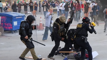 Tres policías antidisturbios con cascos y escudos. Uno con bastón, otro con arma amarilla. Contenedor azul grafitado y personas en el fondo borroso