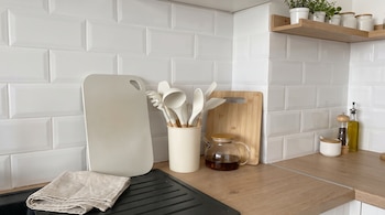 Utensilios blancos, dos tablas de cortar, una tetera de vidrio y frascos sobre una mesada de madera frente a una pared de azulejos.