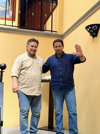 Nery Ramos (camisa azul) y Alfonso Portillo (camisa beige) de pie ante una pared amarilla. Ramos saluda. Detrás, una barandilla y un libro abierto