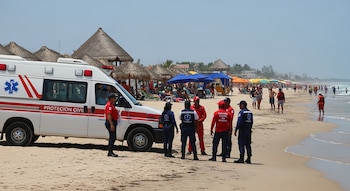Una ambulancia y personal de Protección Civil en una playa con bañistas, sombrillas y palapas. El mar y la arena se extienden al fondo.
