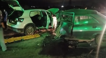 Dos vehículos, uno blanco y uno oscuro, con daños por un choque. Personas y una camilla amarilla se observan en el asfalto durante la noche