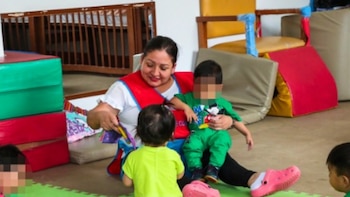 Una mujer sentada en el suelo de un aula de juegos sostiene a un niño en su regazo mientras interactúa con otros pequeños
