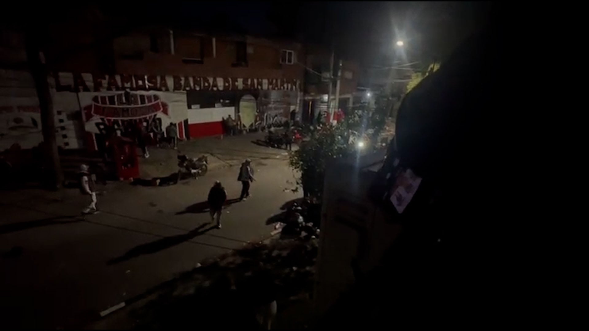 Integrantes de la hinchada de Chacarita Juniors se desplazan por una calle de noche frente a un mural del club durante un cruce interno.