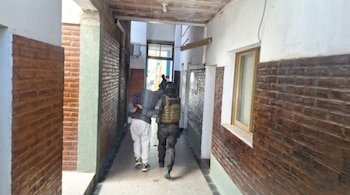 Un hombre esposado con el rostro cubierto es escoltado por un agente de seguridad con casco y chaleco táctico por un pasillo con paredes de ladrillo