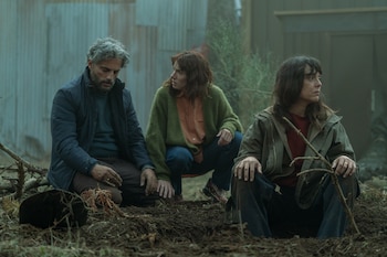 Tres personas, dos mujeres y un hombre, agachadas al aire libre sobre tierra oscura con ramas y hojas, en un entorno con poca luz y fondo borroso