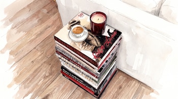 Pila de libros de moda con un libro de Ralph Lauren, una taza de café, una vela roja encendida, piso de madera y el borde de un sofá blanco.
