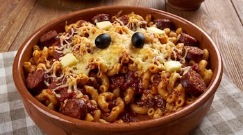 Pasta con salsa de tomate, rodajas de chorizo, queso fundido, mantequilla y dos aceitunas negras dentro de una cazuela de barro sobre un mantel.