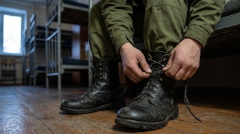 Manos de una persona con pantalón verde que ata los cordones de unas botas negras sobre un suelo de madera en una habitación con literas.