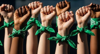 Una fila de puños de mujeres levantados hacia el cielo, con pieles de diferentes tonos, cada muñeca adornada con un pañuelo verde anudado.