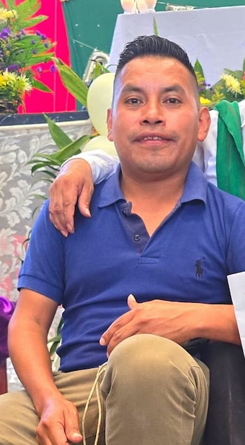 Un hombre con camisa polo azul y pantalón claro posa sentado, mientras la mano de otra persona reposa sobre su hombro