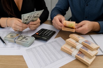 Manos de dos personas contando billetes de dólares y pesos argentinos sobre un escritorio con calculadora y planillas de papel.