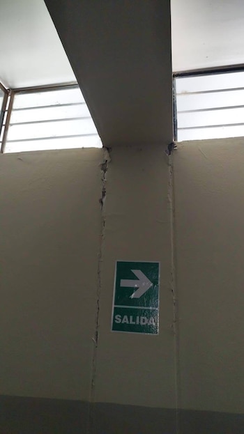 Una pared beige con grietas verticales severas y un letrero verde de 'SALIDA' con una flecha blanca. Arriba, ventanas con barrotes y un techo blanco