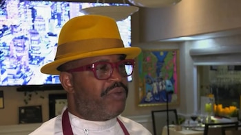 Hombre con sombrero fedora amarillo, gafas rojas, barba, bigote y uniforme de chef. Pantalla de televisión con ciudad, pintura colorida y mesas de restaurante al fondo