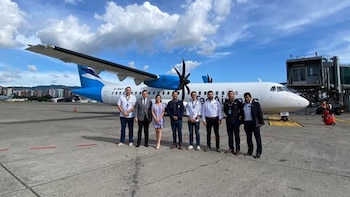 Un grupo de ocho personas, siete hombres y una mujer, de pie en un aeropuerto frente a un avión de hélices blanco y azul de Tag Airlines bajo un cielo nublado