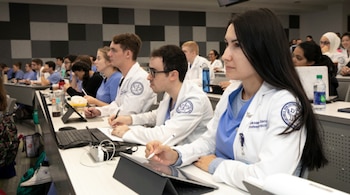Primer plano de varios estudiantes de medicina osteopática con batas blancas, tomando notas en tabletas y cuadernos durante una clase en un aula moderna