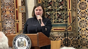 Una mujer con cabello corto y oscuro habla por un micrófono en un podio con el sello de la Asamblea de Nueva York, contra un fondo de paneles decorados