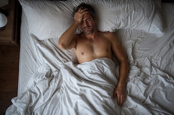Vista superior de un hombre acostado en una cama con sábanas blancas, sin camiseta y con la mano en la frente.