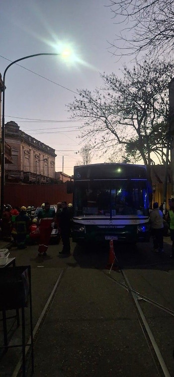 Un colectivo oscuro en una calle con vías de tren, rodeado de personal de emergencia, personas y un cono de señalización en primer plano