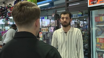 Dos hombres en el interior de una tienda de conveniencia. Uno, con barba y camisa a rayas, mira al frente. El otro está de espaldas a la cámara