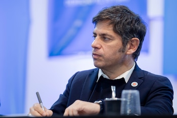 Axel Kicillof, con traje oscuro, camisa y suéter, escribe con un bolígrafo. Un pin redondo en su solapa. Vaso y fondo azul difuminados