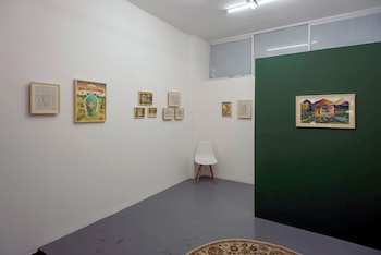 Paredes blancas y una pared verde en galería, nueve obras enmarcadas, una silla blanca, parte de una alfombra, ventanas altas y una luz de techo