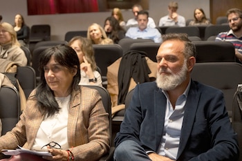Un hombre con barba blanca y una mujer con saco a cuadros están sentados en las butacas de un auditorio