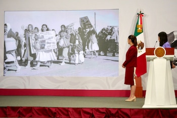 Mujer de espalda con traje rojo frente a una pantalla con foto en blanco y negro de una protesta de mujeres. A su lado, un podio con el escudo de México y la bandera