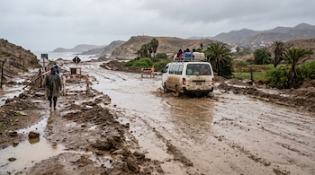 Una furgoneta blanca con personas en el techo transita por un camino lodoso e inundado cerca de un peatón con poncho.