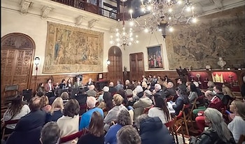 Público sentado de espaldas frente a una mesa con ponentes en una sala con tapices en los muros y lámparas de araña
