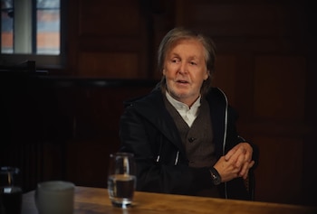 Paul McCartney, con cabello gris y barba, sentado en una mesa de madera oscura. Viste una chaqueta negra, chaleco marrón y camisa blanca, con un vaso de agua