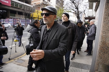 Hombre con gorra de cuero, gafas de sol y traje a rayas camina en una acera, rodeado de camarógrafos y otras personas. Al fondo, edificios y árboles