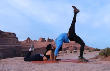 Hombre practicando yoga en un tapete azul junto a un río verde. Viste camiseta beige y shorts azules. Al fondo, edificios coloridos y montañas