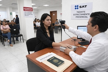 Un empleado registra datos biométricos de una mujer en una oficina del SAT. Se ven un escáner, documentos de identidad y otras personas esperando.