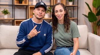 Hombre y mujer sentados en un sofá de tela beige. El hombre lleva gorra negra, sudadera azul, cadena de plata y reloj dorado. La mujer usa camiseta verde y jeans.