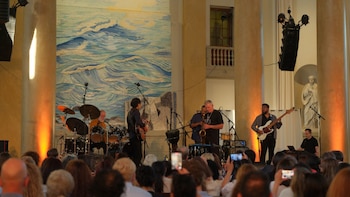 Un quinteto musical actúa en un escenario iluminado frente a una audiencia. Detrás hay un tapiz de olas, columnas y una estatua. El público graba con teléfonos