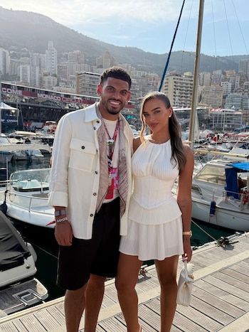 Morgan Gibbs-White y una mujer vestida de blanco posan sonrientes en un muelle con yates y edificios al fondo