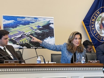Una mujer frente a un micrófono señala un cartel con la imagen de una pista de aeropuerto en un salón de audiencias