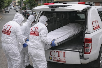 Tres agentes del CTI con trajes blancos y tapabocas introducen un cadáver cubierto en una camilla al compartimiento trasero de un vehículo oficial blanco.