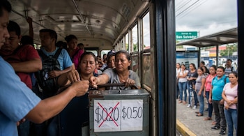 Personas pagando en un autobús lleno, donde un cartel indica el aumento del pasaje de $0.20 a $0.50, con pasajeros esperando en una parada exterior.
